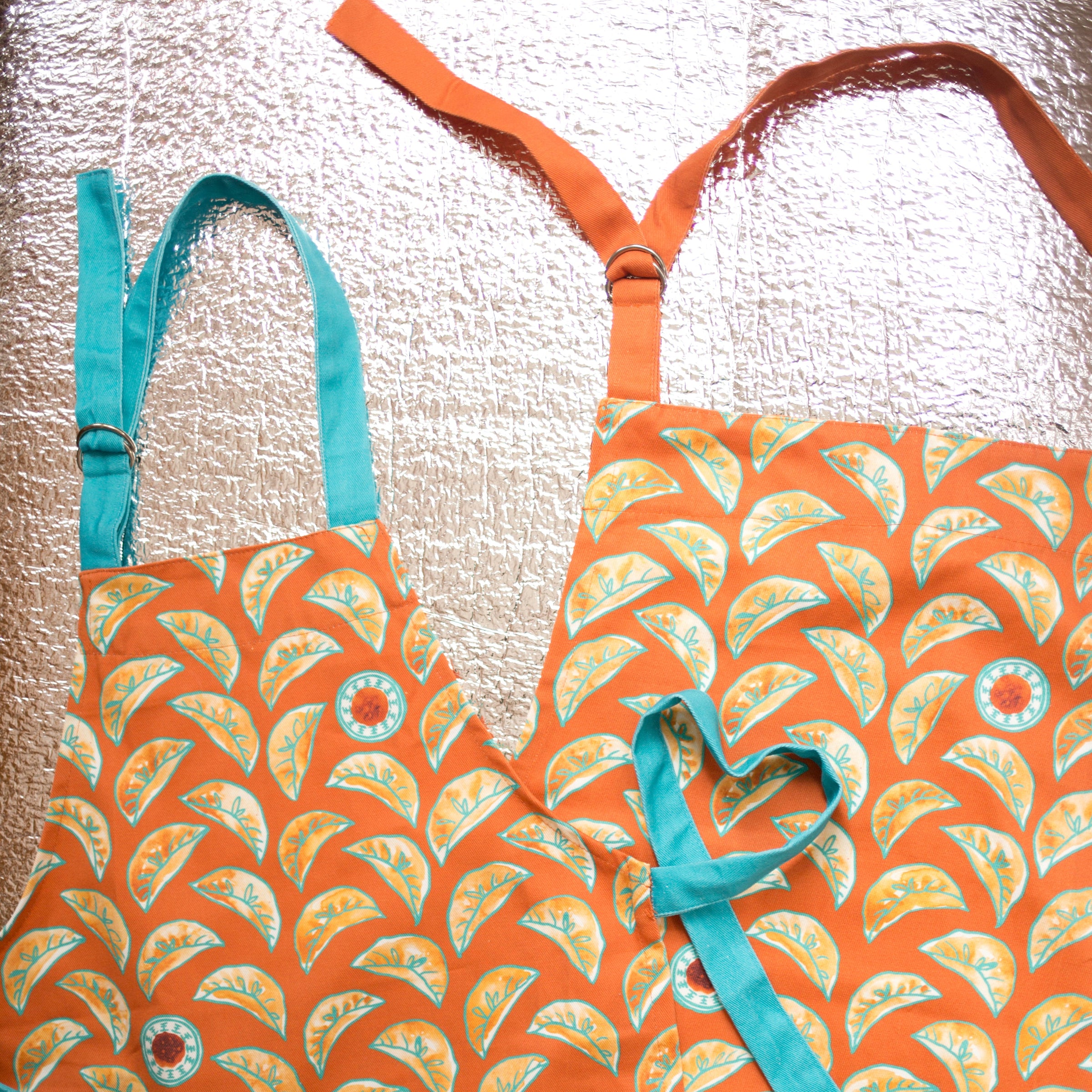 kid's dumpling apron with teal straps, shown left of an adult-sized one with orange straps